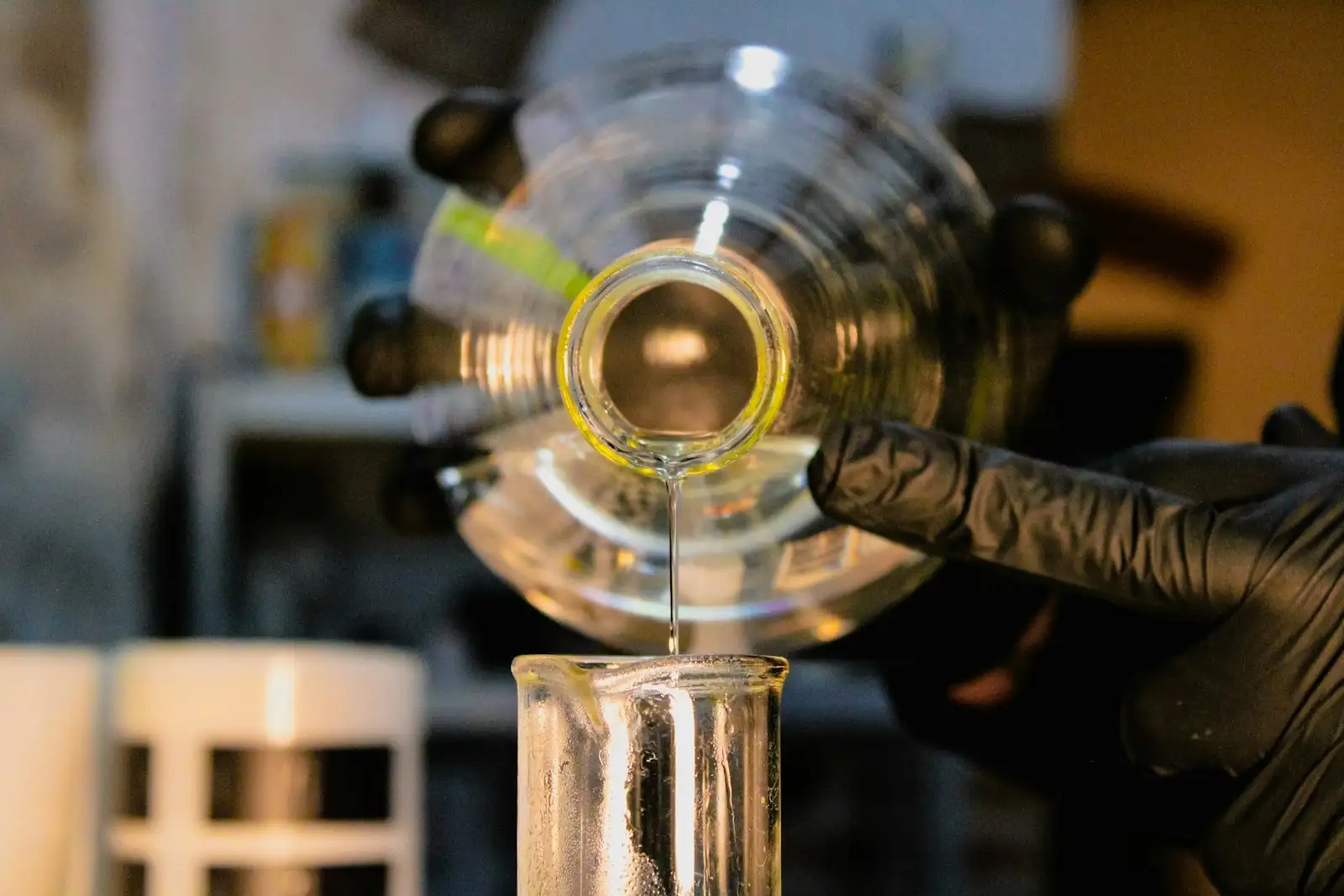 Gloved hand pouring a liquid into a beaker in a dimly lit chemistry lab