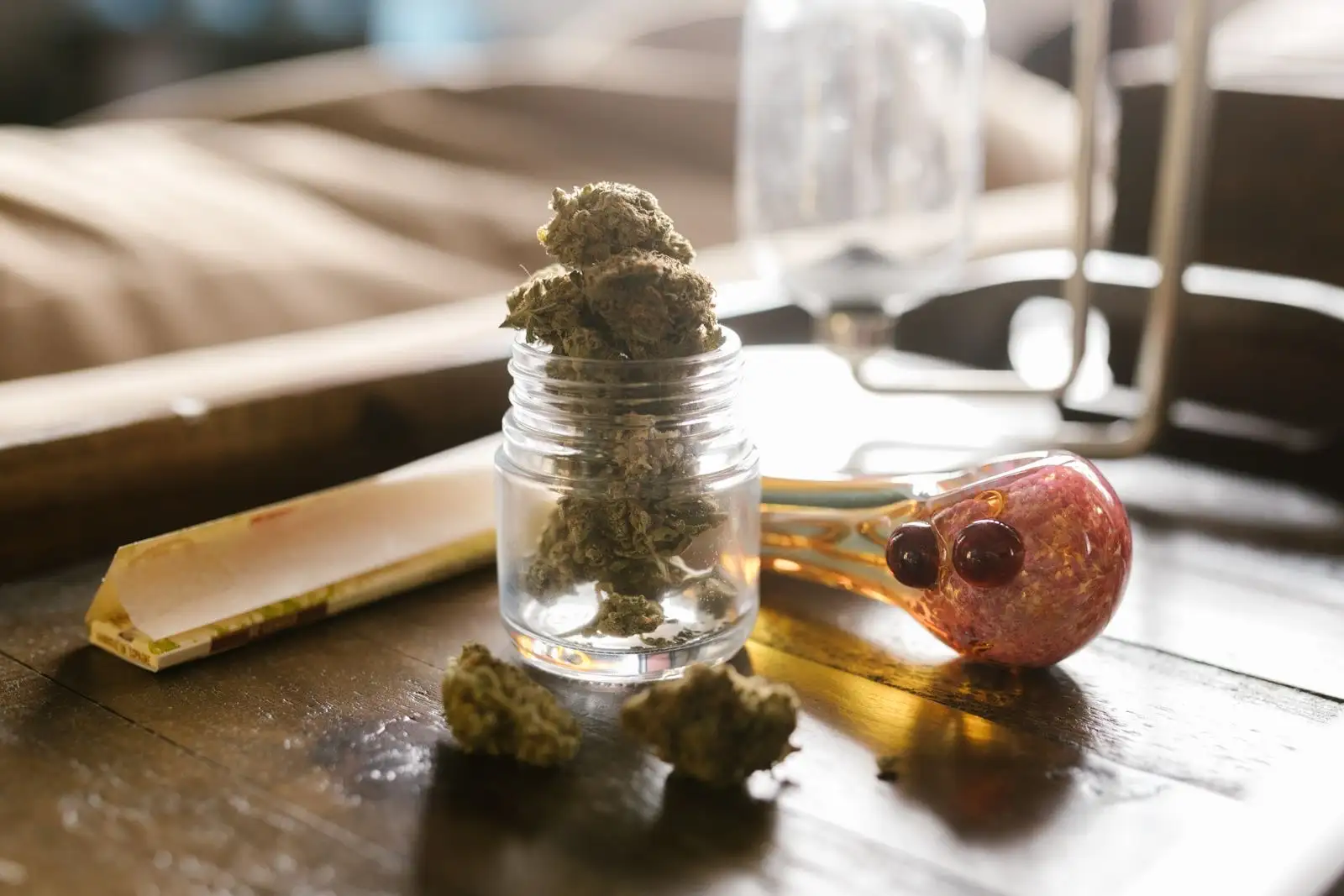 Small glass jar filled with dried hemp buds on a wooden table, next to a pack of rolling papers