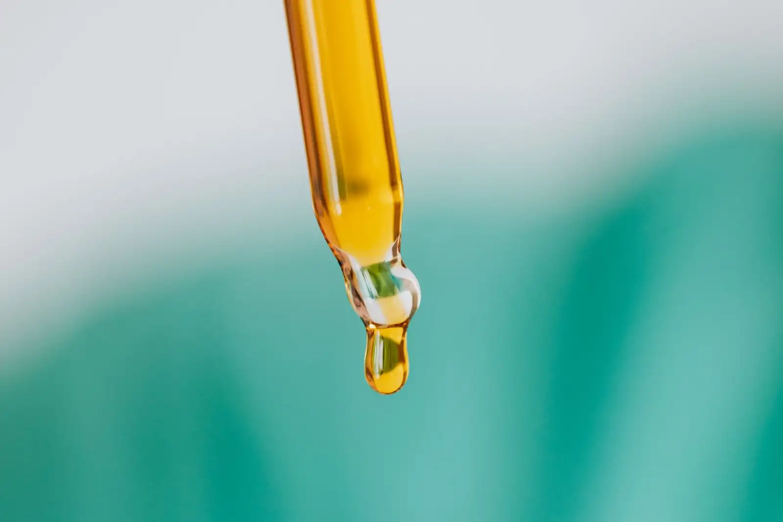 Glass pipette releasing a drop of amber oil, against a blurred green background