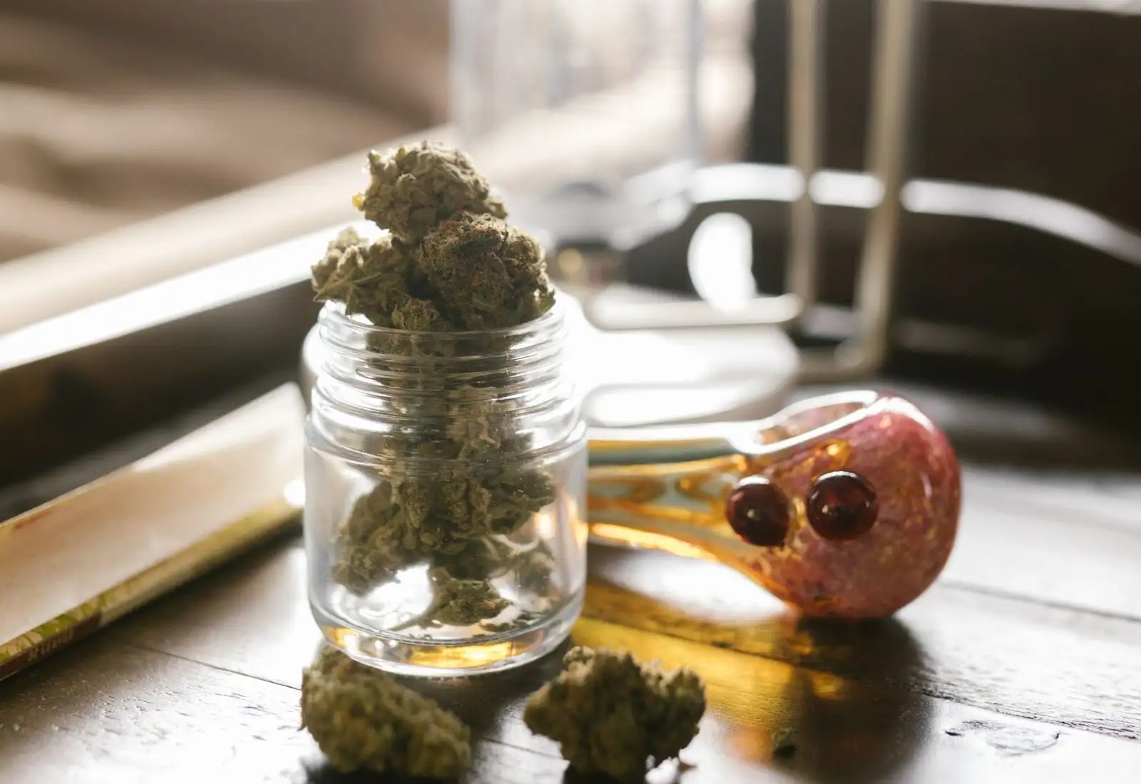 Glass jar filled with dried hemp flowers, next to a glass pipe and a booklet of rolling papers