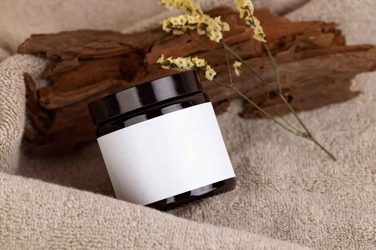 Black cosmetic jar with a blank white label, resting on a towel next to a piece of bark