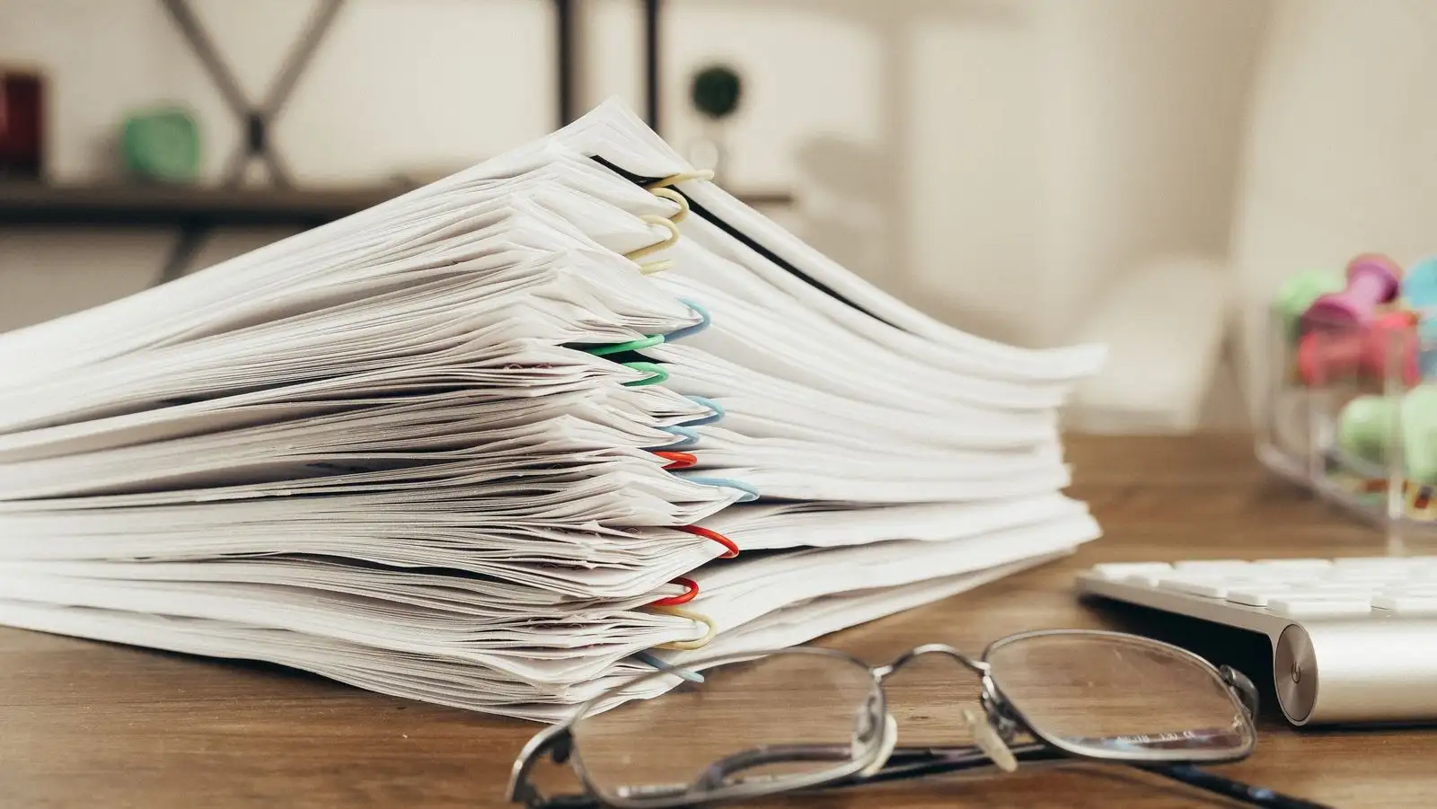 A stack of clipped documents on a desk, next to a pair of glasses