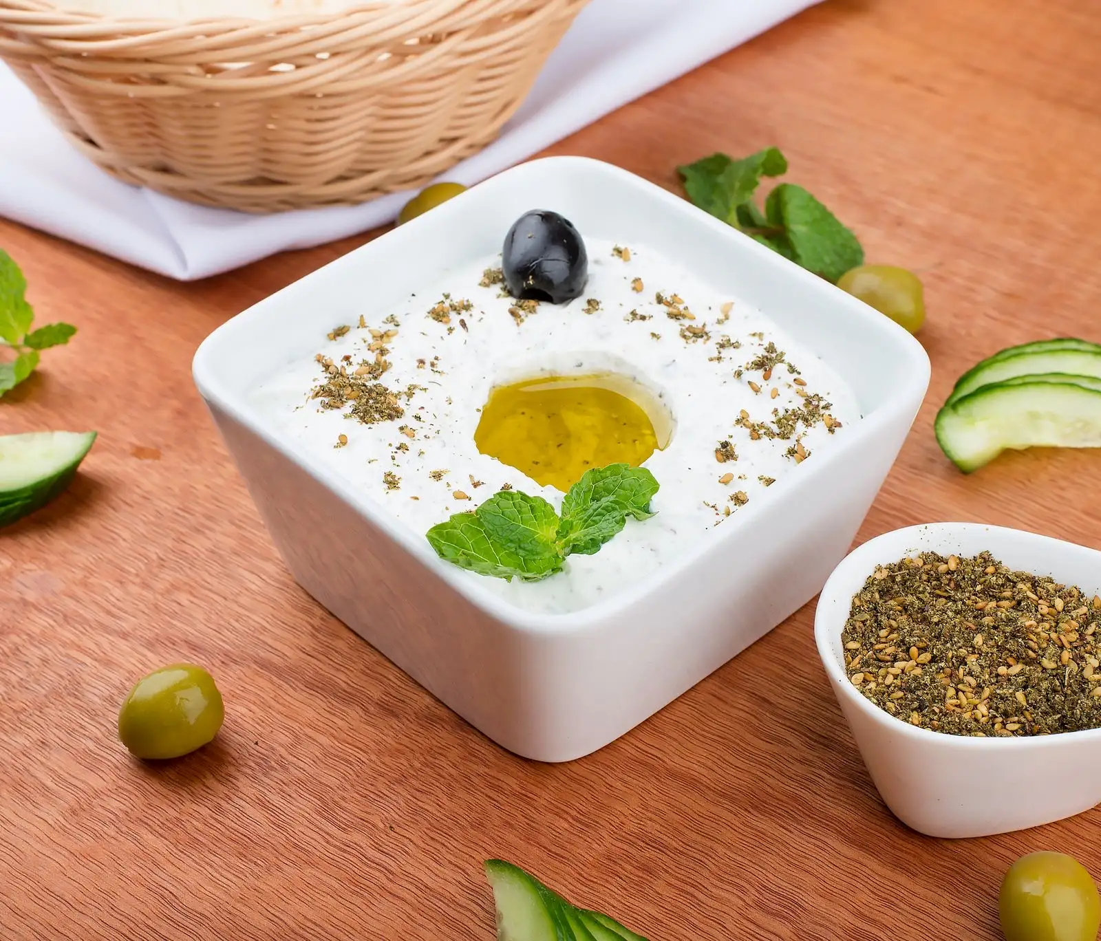 Tzatziki in a square white bowl, drizzled with olive oil and topped with mint, surrounded by cucumber slices and olives