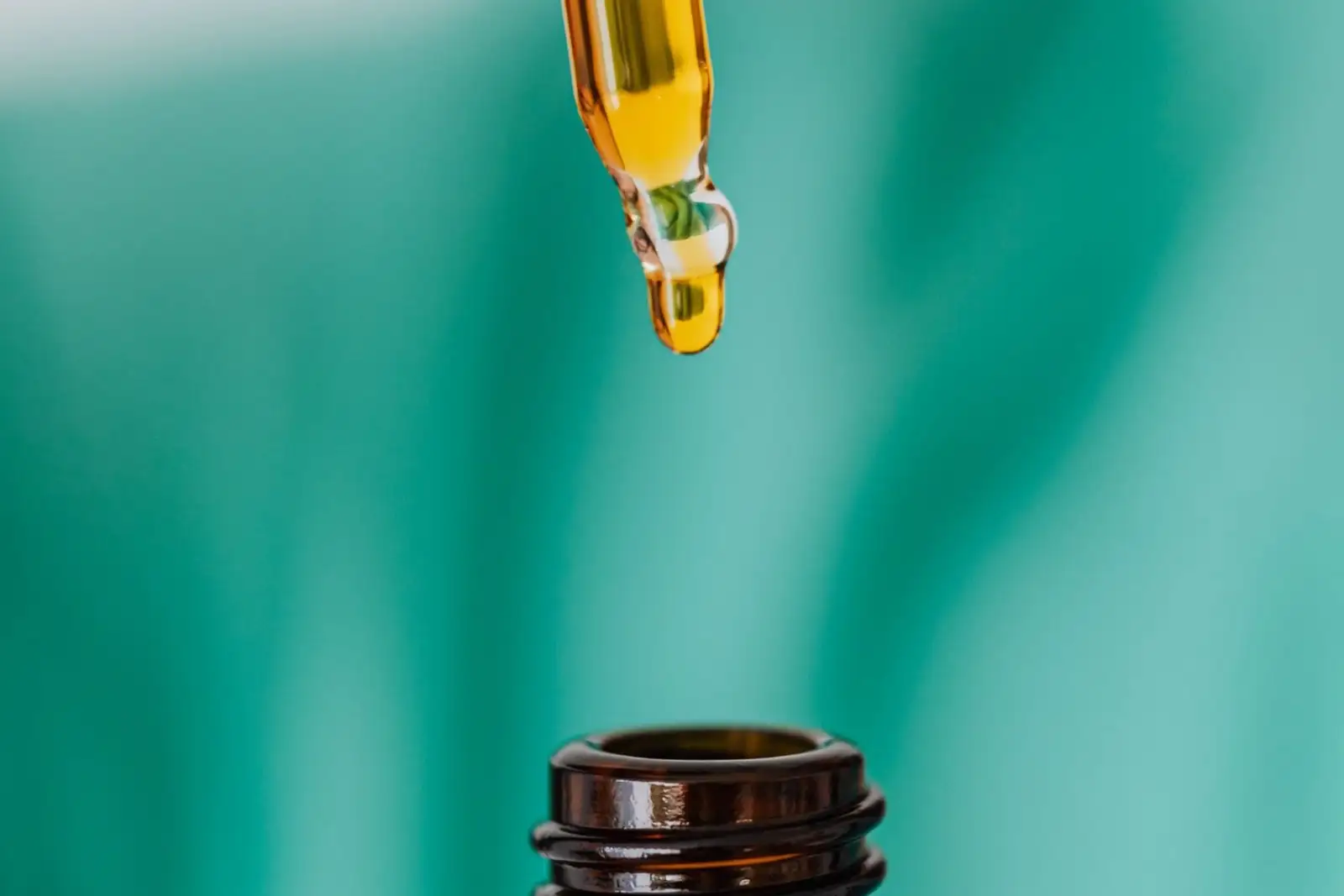 A drop of golden oil at the tip of a glass pipette, above an amber bottle