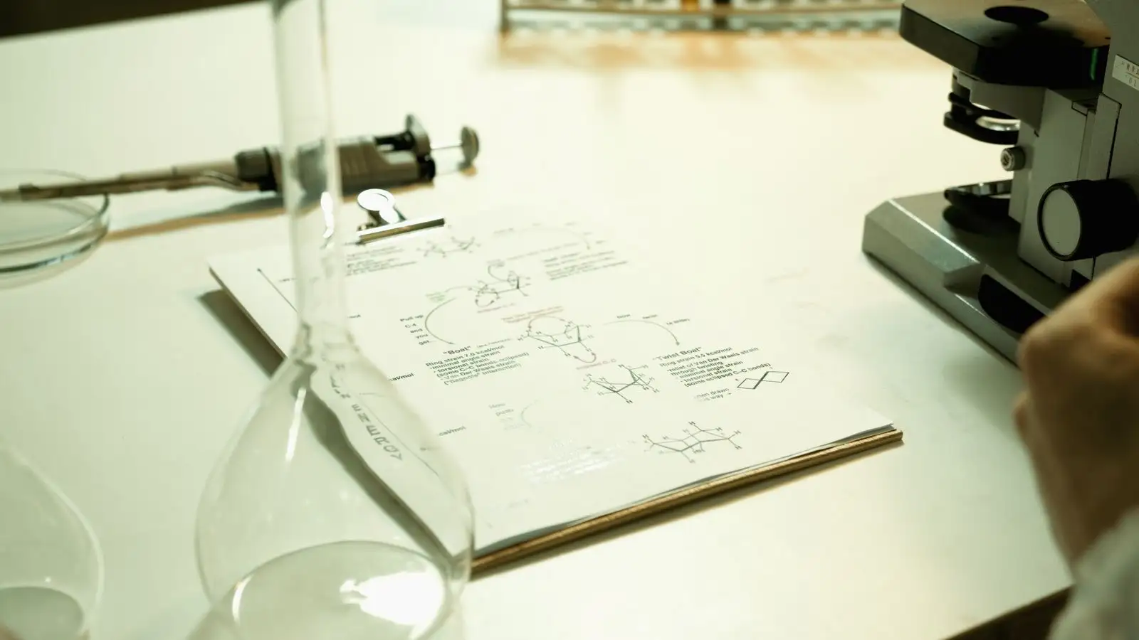 Laboratory bench with a sheet of chemical structures on a clipboard, a glass flask and a microscope