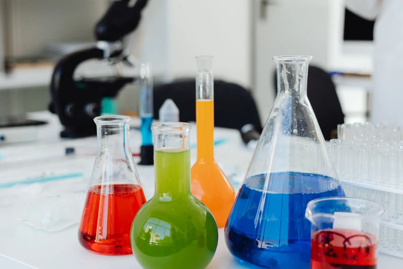 Laboratory glassware on a bench, flasks filled with coloured liquids in front of a microscope