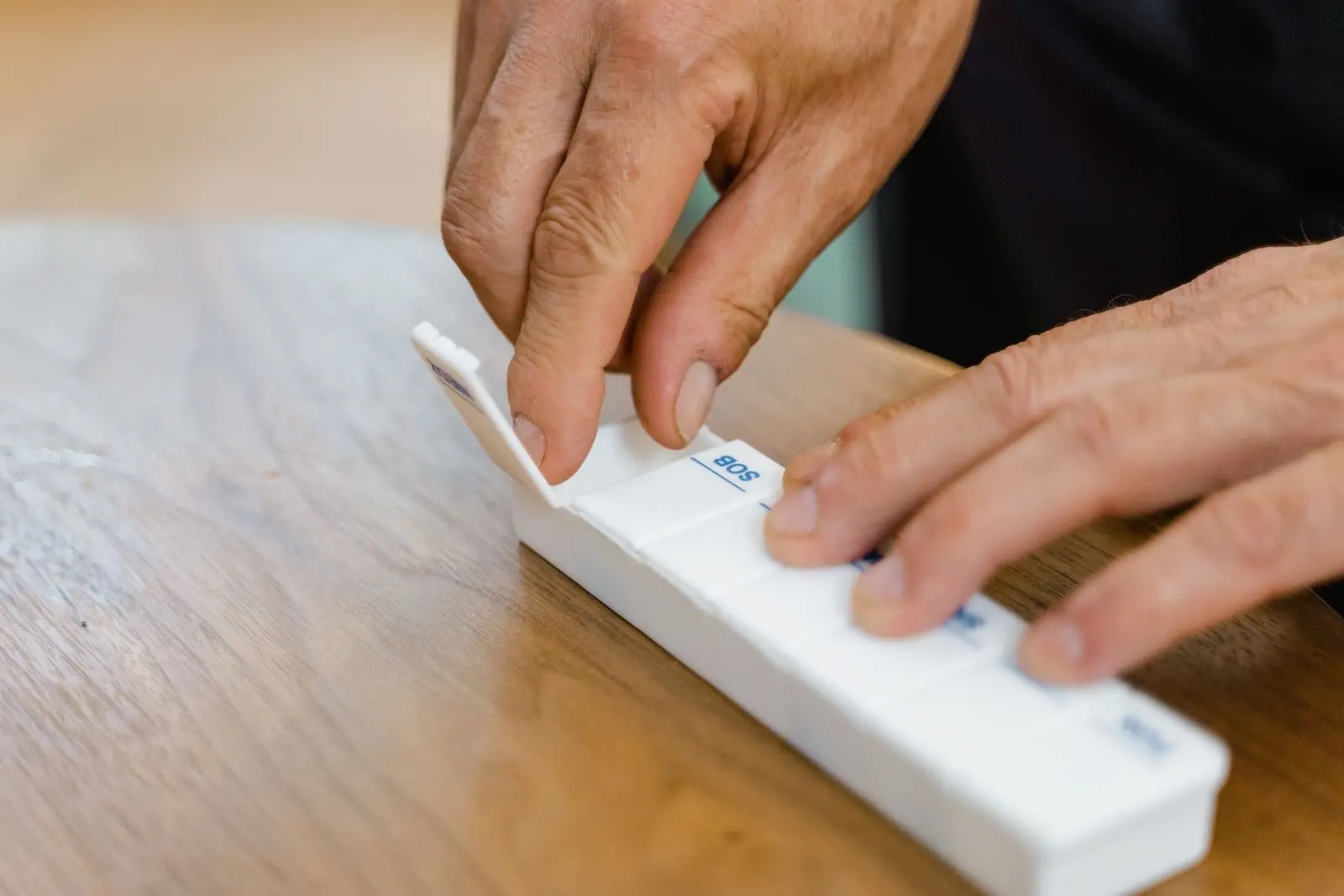 Hands of an older person sorting medicines into a weekly pill organiser on a wooden table
