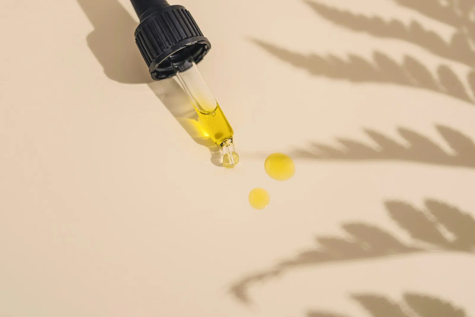Dropper pipette filled with golden oil, two drops resting on a beige background
