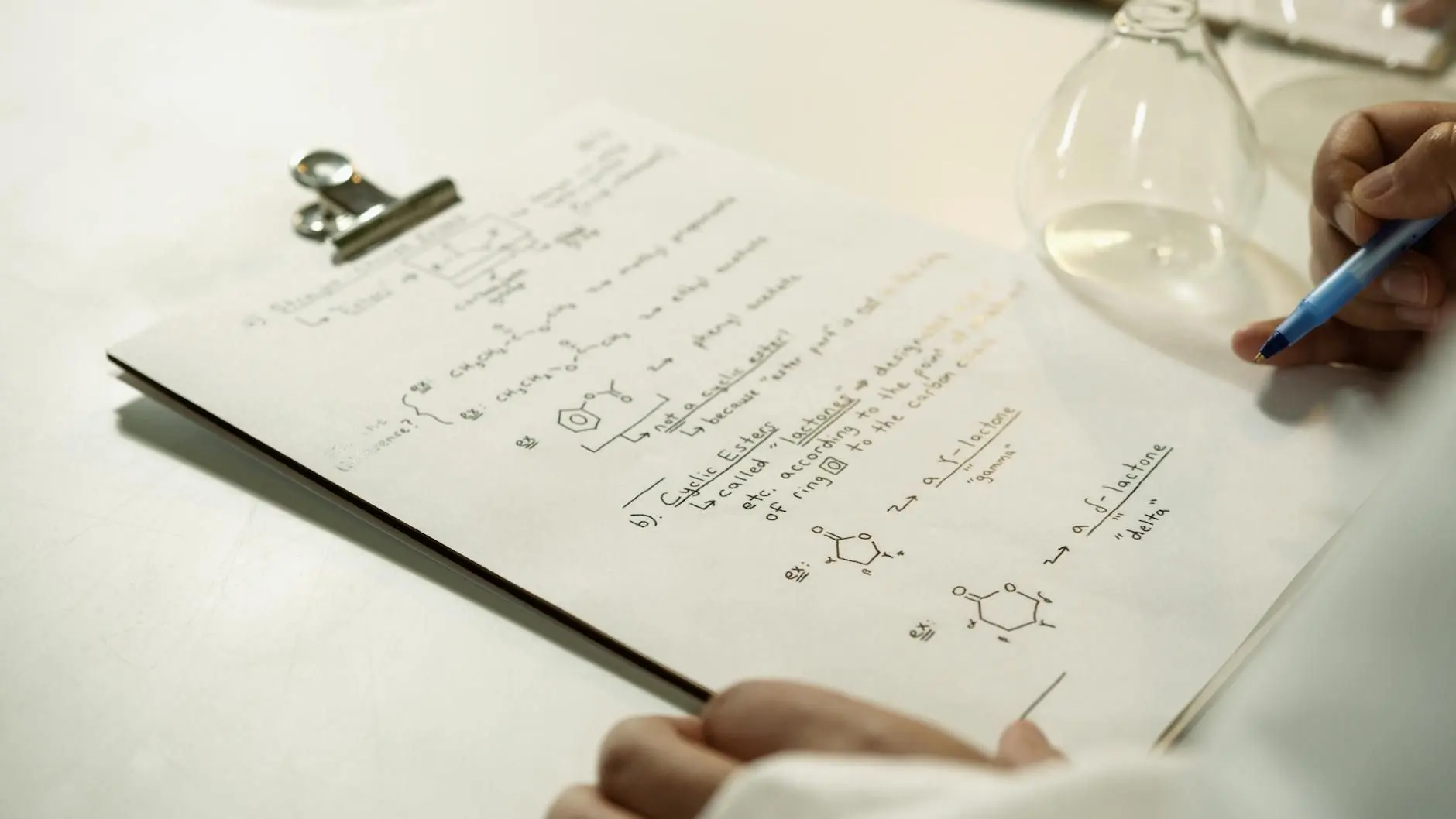Handwritten organic chemistry notes on a clipboard, next to laboratory glassware