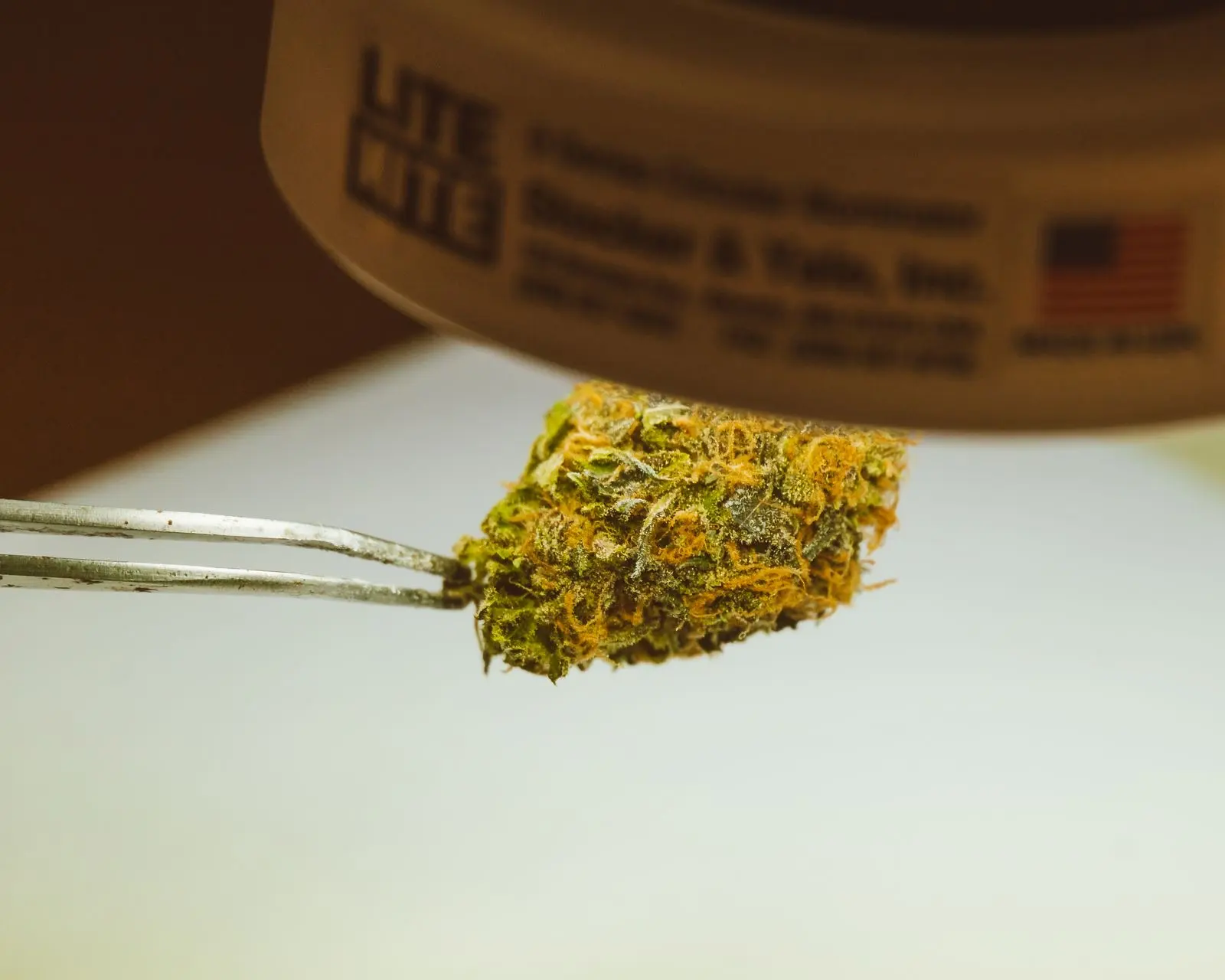 Dried hemp bud held with tweezers under a lamp, the careful gesture of loading a chamber